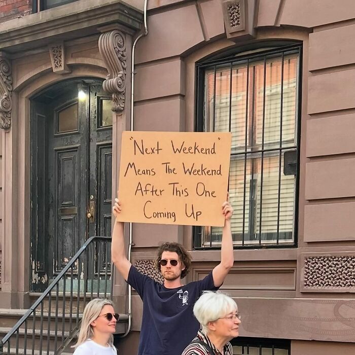 Dude With The Sign holding a cardboard sign about the meaning of next weekend outside a brownstone building.