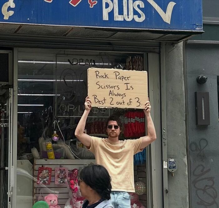 Man wearing sunglasses holding a sign about rock paper scissors in a humorous dude with the sign street photo.