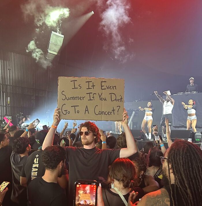 Man with the sign at a crowded concert holding cardboard text reflecting thoughts everyone is thinking in a live music setting.