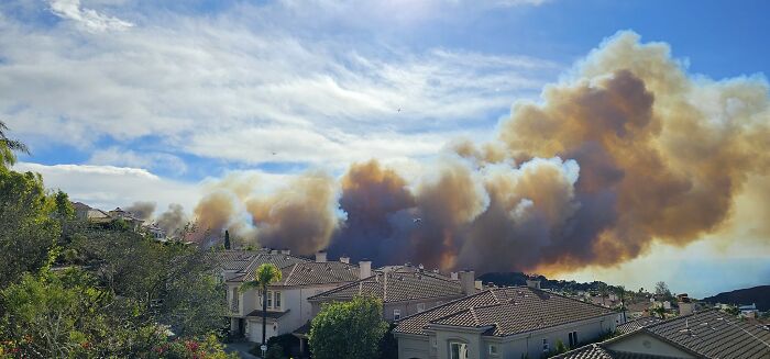 Thick smoke from a catastrophic wildfire billows over residential homes under a partly cloudy sky in a recent wildfire event.