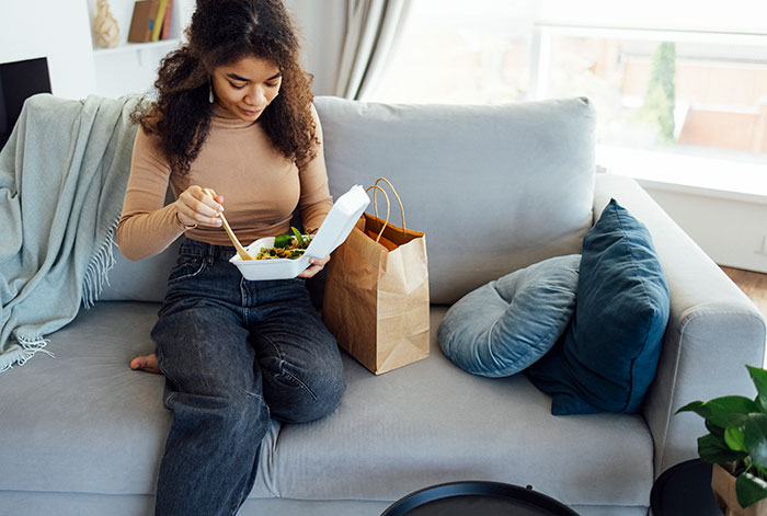 Young woman sitting on a couch eating DoorDash takeout while babysitting, showing order Doordash while babysitting overreaction. Young woman sitting on a couch eating DoorDash takeout while babysitting, showing order Doordash while babysitting overreaction.
