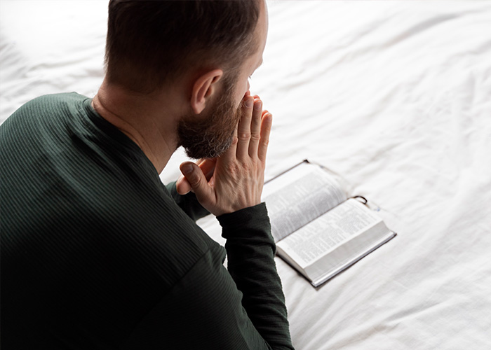 Man in dark shirt sitting on bed with hands together near mouth, looking thoughtfully at an open book or Bible.
