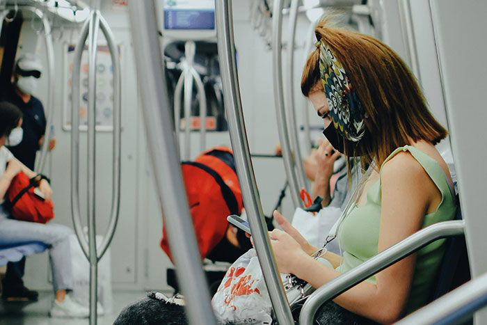 Woman wearing a mask and patterned hair scarf sits on public transit, using phone, reflecting unhinged microfeminisms in a candid moment.
