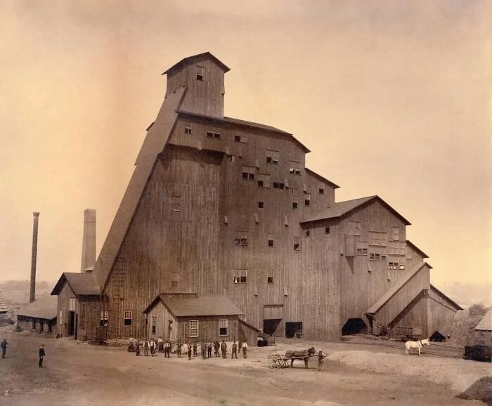 Large historic wooden industrial building with workers and horses in front, showcasing old architecture.