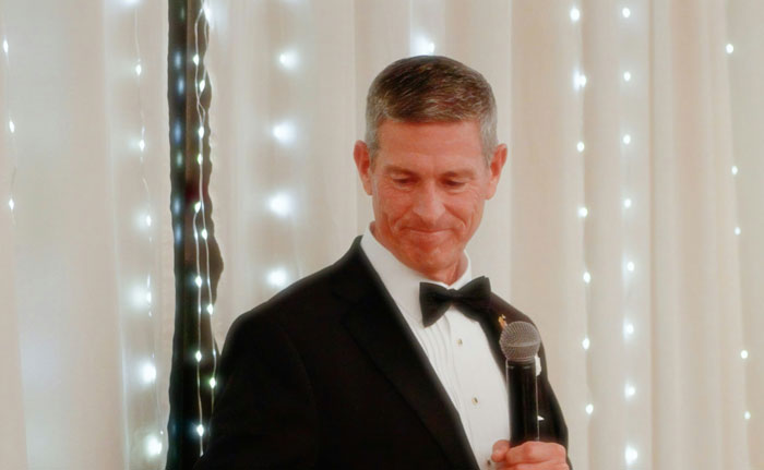 Man in a black tuxedo holding a microphone, standing in front of wedding decor with string lights. - 6