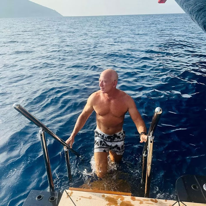 Jeff Bezos showing new look with hair, climbing out of the ocean onto a boat on a sunny day.