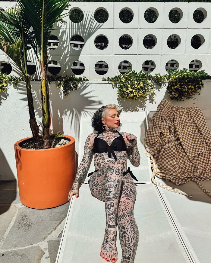 Woman with full body suit tattoos relaxing outdoors on a lounge chair beside a large orange planter and plants.