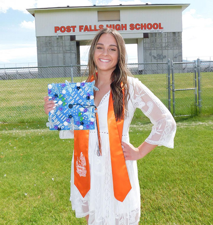 Graduate in white dress and orange sash holding decorated cap in front of Post Falls High School, unrelated to unsealed docs or crime details. - 8