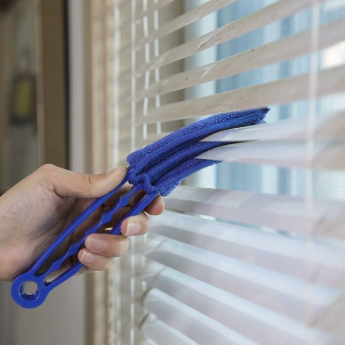 Hand using a blue cleaning tool to dust window blinds, illustrating solutions for annoying roommate problems.