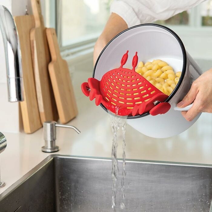 Red crab-shaped kitchen invention strainer draining water from pot with pasta over a sink.