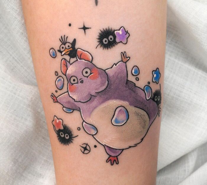 Colorful cartoon-style tattoo of a whimsical purple creature with bubbles and cute black soot sprites on skin.