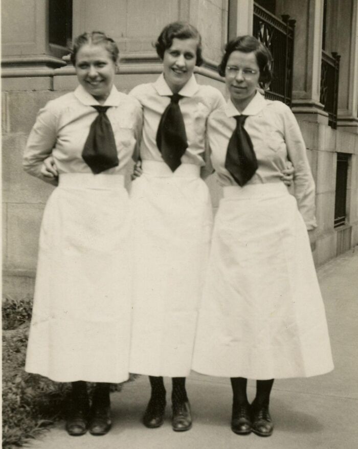Three vintage nurses standing arm in arm outside a building, wearing traditional uniforms and ties, smiling at the camera