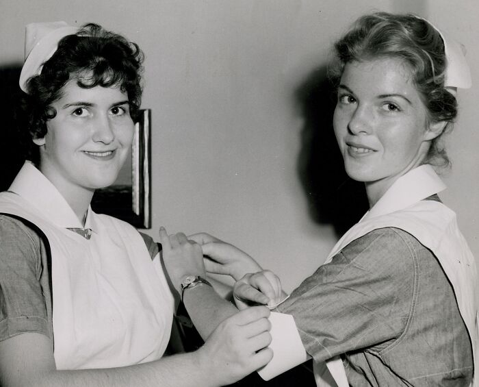 Two vintage nurses smiling while preparing an arm bandage, showcasing classic nursing uniforms and care.