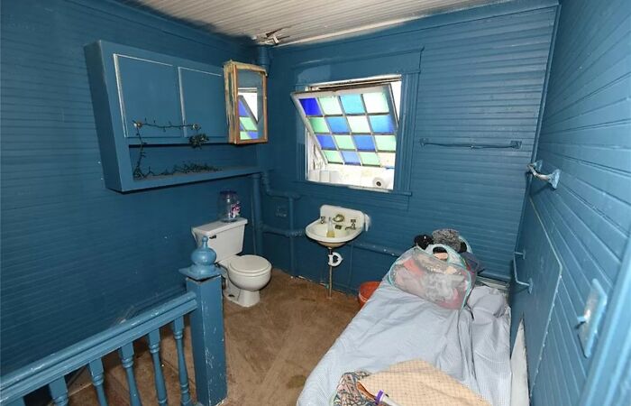 Small cramped bathroom painted blue with toilet, tiny sink, stained carpet, and a bed, showing unhinged real estate listings.