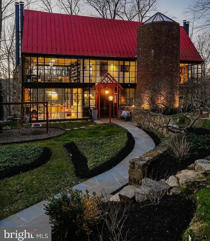 Unhinged real estate listing featuring a unique glass house with a red roof and a large brick silo at dusk.