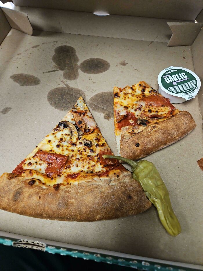 Two uneven pizza slices with a pepper placed in a greasy pizza box, an epic work fail in food delivery presentation.