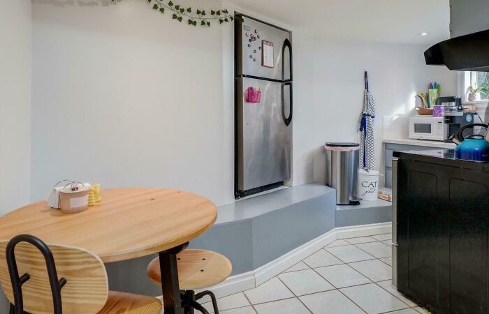 Small kitchen with an elevated fridge area, round wooden table, and mismatched decor in an unhinged real estate listing.