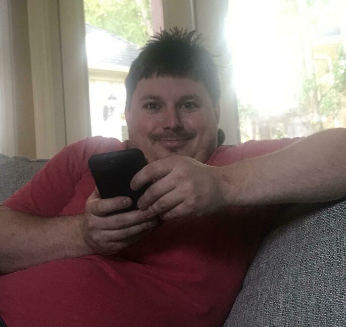 Man in red shirt holding phone and smiling while lounging on a couch, representing salon glow-up and breakdown stories.