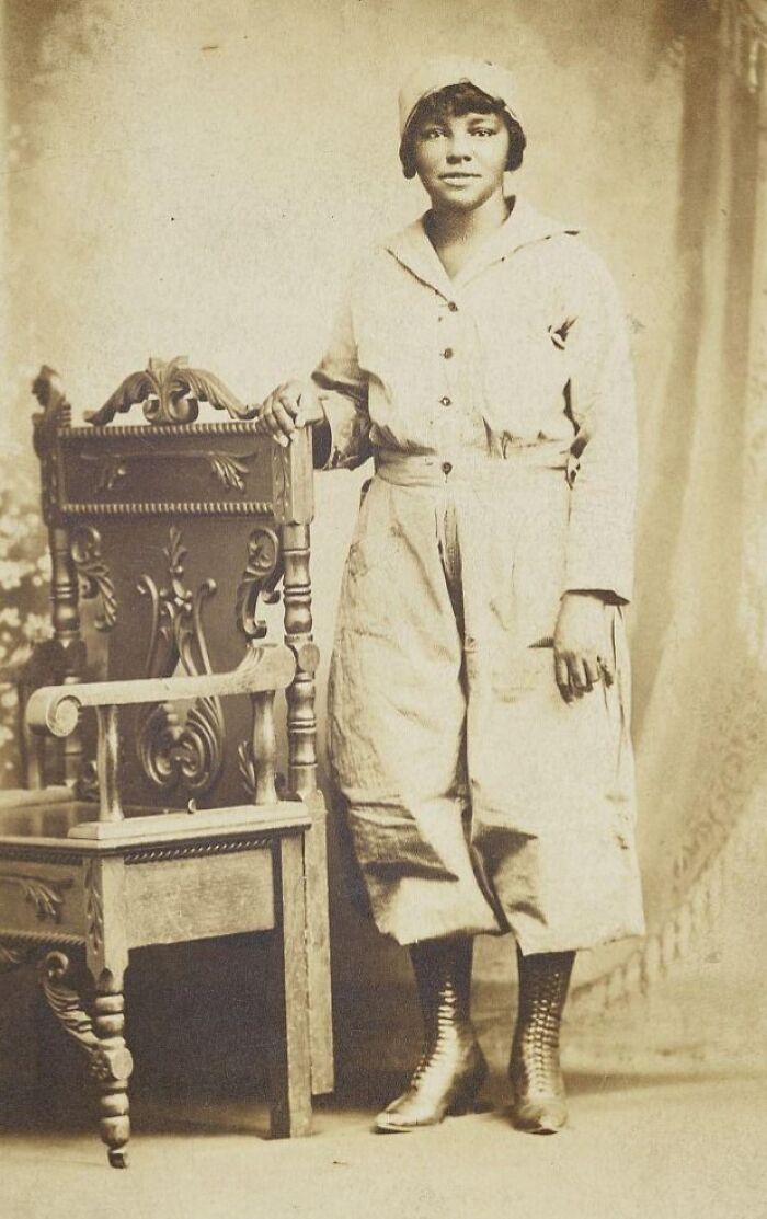 Young woman in vintage outfit posing beside an ornate chair in a classic 1930s vintage postcard style photograph