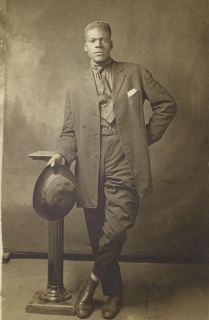 Vintage postcard shows a stylish young man from the 1930s leaning on a pedestal holding a hat, wearing a suit and tie.