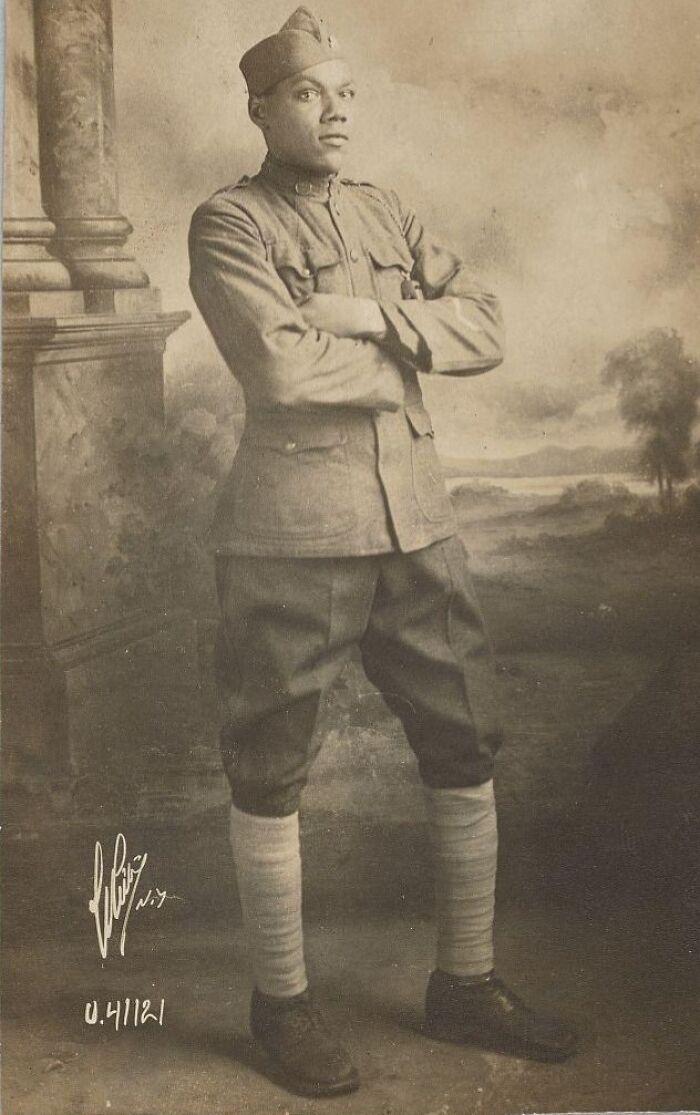 Vintage postcard showing a confident 1930s soldier in uniform standing with arms crossed against a classic backdrop.