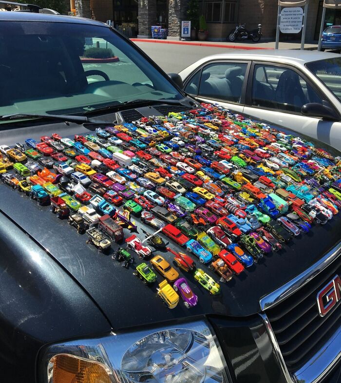 Toy cars glued to the hood of a black car as an outrageous car mod in a parking lot on a sunny day