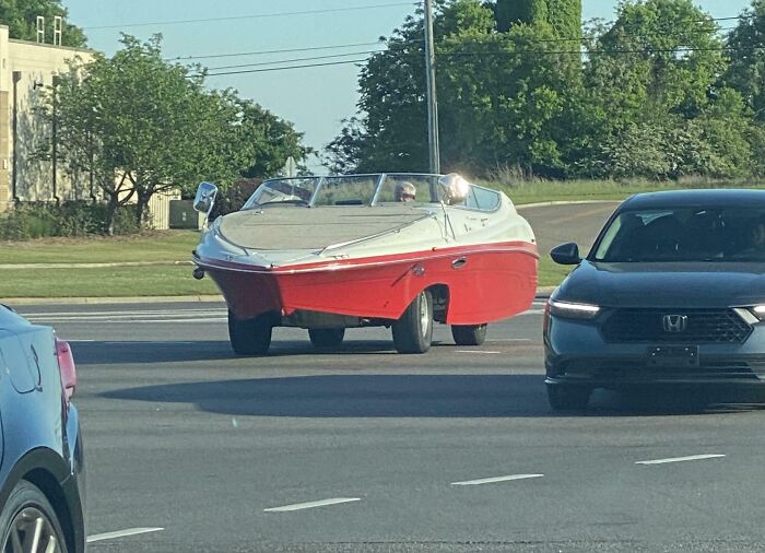 Red and white car modified to look like a boat driving on a city street among other vehicles, car mods visible
