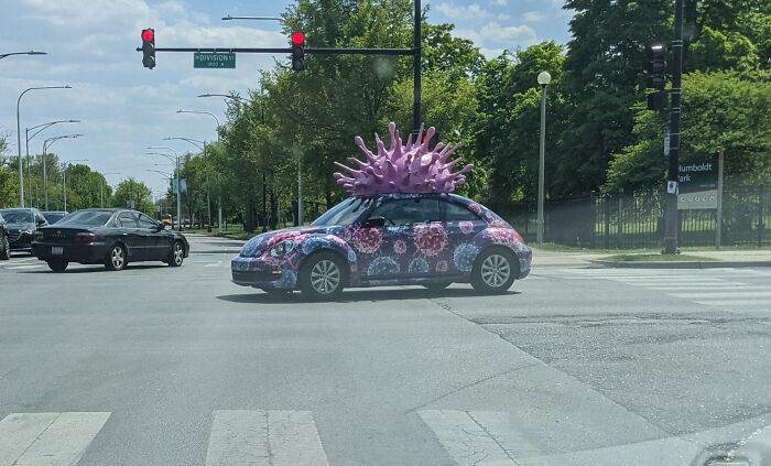 Car covered in virus-themed paint with large pink spiky mod on roof, an example of outrageous car mods on city street.