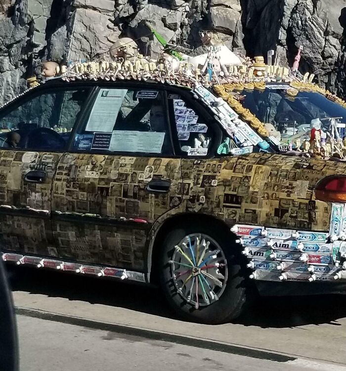 Car covered with photos, dolls, dental products, and various items, showcasing outrageous car mods on a city street.