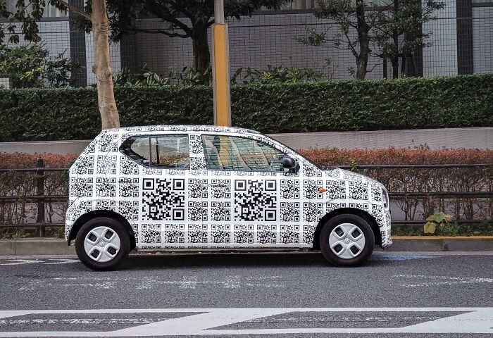 Small car covered entirely in black and white QR code car mods, parked next to sidewalk with trees and bushes behind.