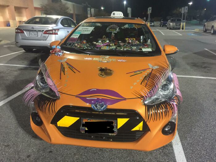 Orange car with outrageous modifications, including large eyelashes, lips decal, and various colorful decorations on the hood and windshield.