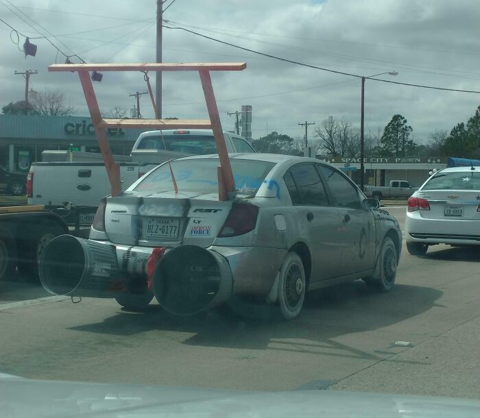 Small silver car with outrageous car mods including large pipes on the back and a wooden spoiler frame on top.
