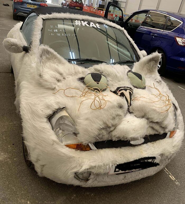 Car covered in white fur with exaggerated cat face features as an outrageous car mod parked in a garage.