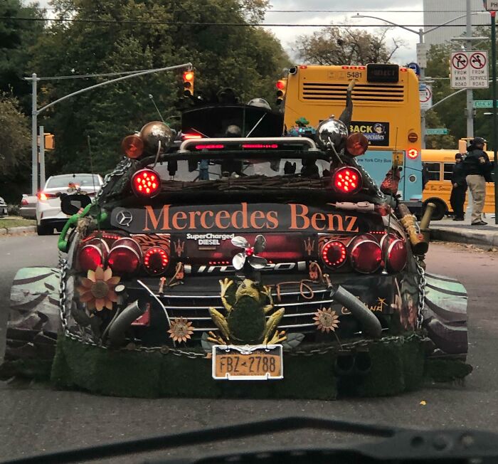 Outrageous car mods on a Mercedes Benz with oversized rear lights, decorative frog, and floral accents on a city street.
