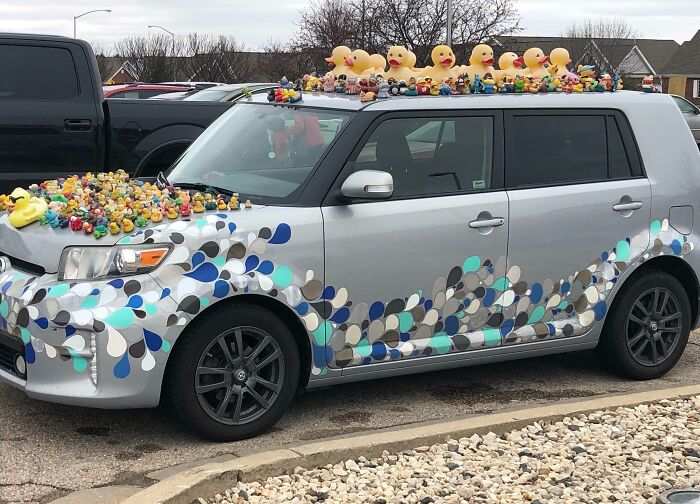 Silver car with colorful teardrop decals and numerous rubber duck car mods covering the hood and roof in a parking lot.