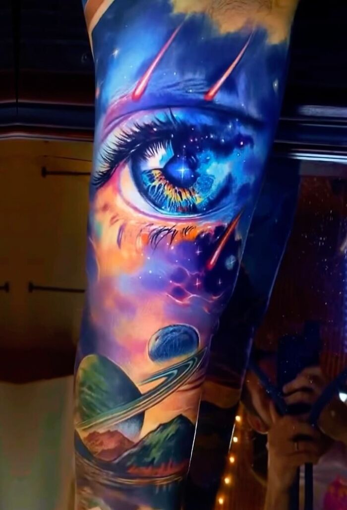 Vibrant 3D tattoo featuring a cosmic eye, planets, and meteors, creating a portal to another world on a forearm.