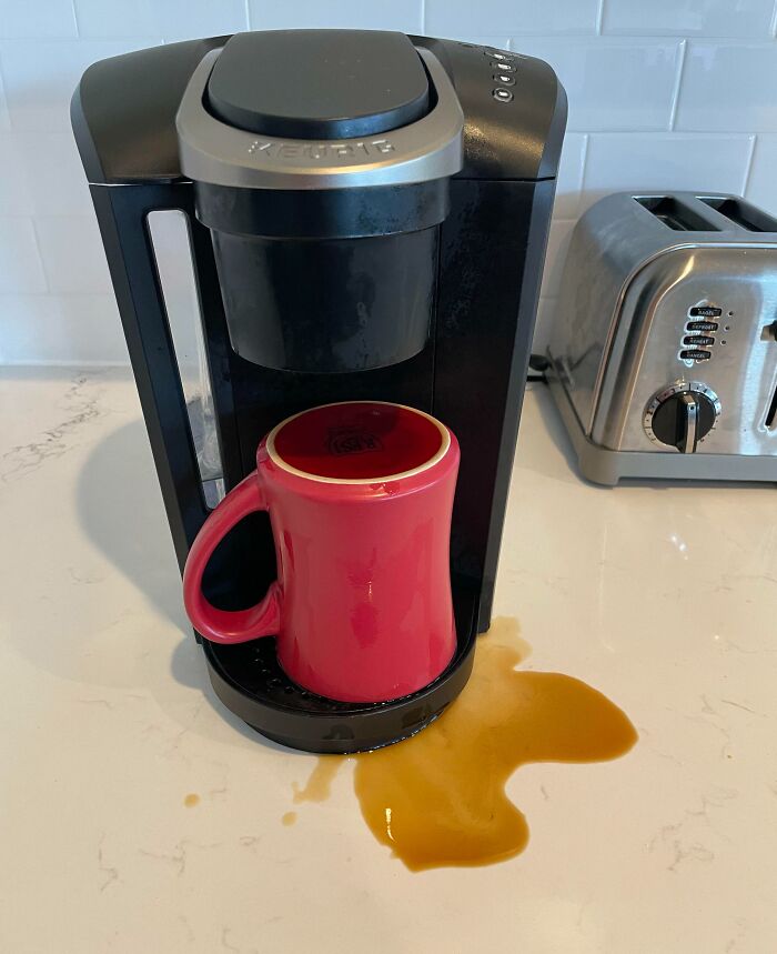 Coffee maker with a red mug placed incorrectly, causing coffee to spill on the counter, showing clueless partner mishap.