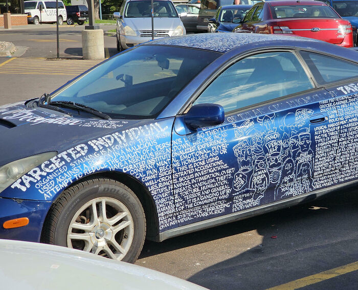 Blue car covered in outrageous custom text and illustrations as part of extreme car mods in a parking lot.