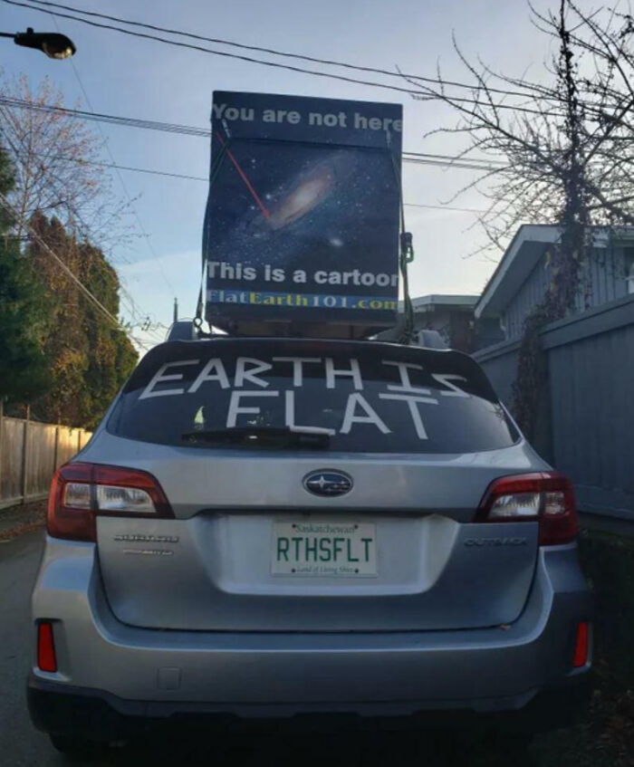Silver Subaru Outback with outrageous car mods, featuring flat earth signs and customized license plate in a residential area.