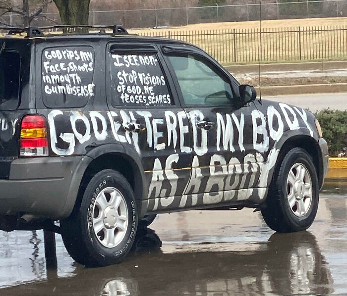 SUV with outrageous car mods featuring large, hand-painted white text covering the vehicle’s exterior on a rainy day.