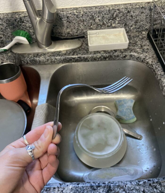 Bent fork held over a kitchen sink full of dirty dishes, illustrating clueless partner humor in a domestic setting.