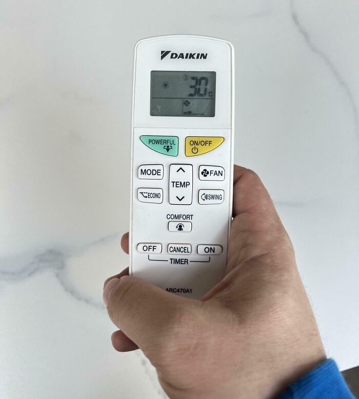 Hand holding a Daikin remote control set to 30 degrees Celsius, illustrating clueless partners with appliances.