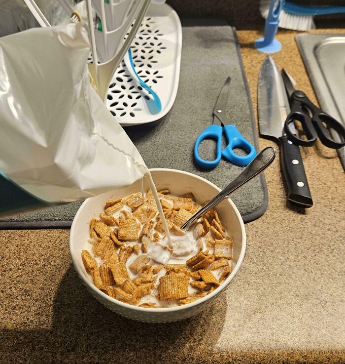 Pouring milk into a bowl of cereal with kitchen scissors and knives in the background showing clueless partner humor.