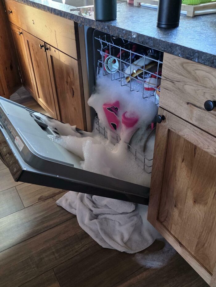 Dishwasher overflowing with soap suds onto wooden kitchen floor, illustrating clueless partner mishap in a humorous home setting.