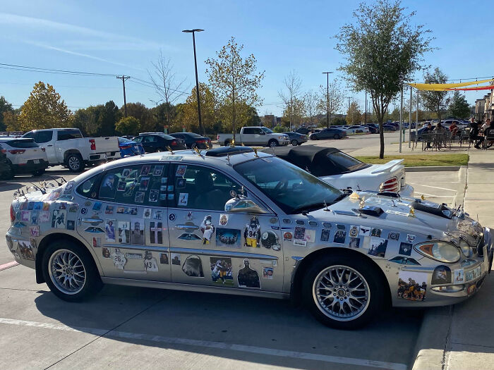 Silver car with outrageous car mods covered in photos and small figurines parked in a busy outdoor lot on a sunny day.