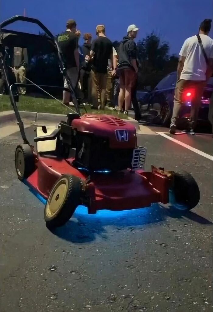 Red lawn mower with blue underglow lighting parked on pavement at night with group of people nearby smiling and chatting.