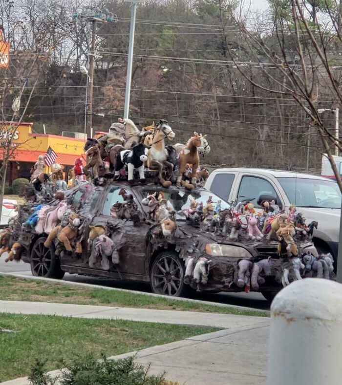 Car covered in numerous horse stuffed animals as an outrageous car mod parked on a street with trees and other cars nearby.