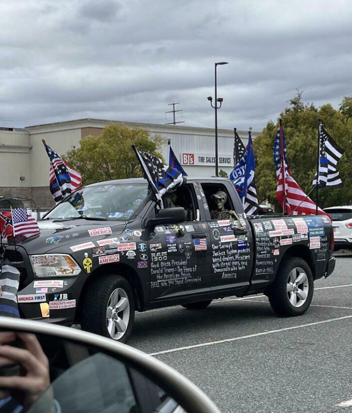 Black pickup truck covered in numerous stickers and mounted flags showcasing outrageous car mods and personalized decorations.