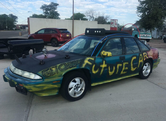 A car with outrageous mods featuring colorful graffiti, rooftop attachments, and custom side mirrors in a parking lot.