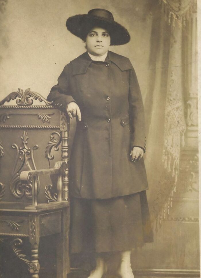 Vintage postcard showing a woman in 1930s attire standing next to an ornate wooden chair.
